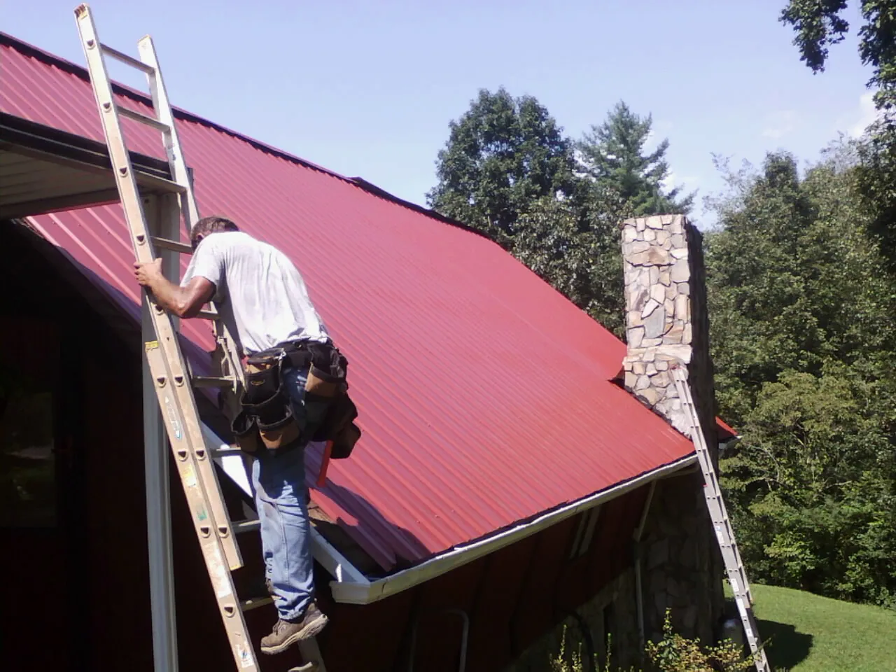 Roofer on a ladder beside a red metal roof and stone chimney — Soffit & Fascia Repair by Roofing Brothers in West Roxbury